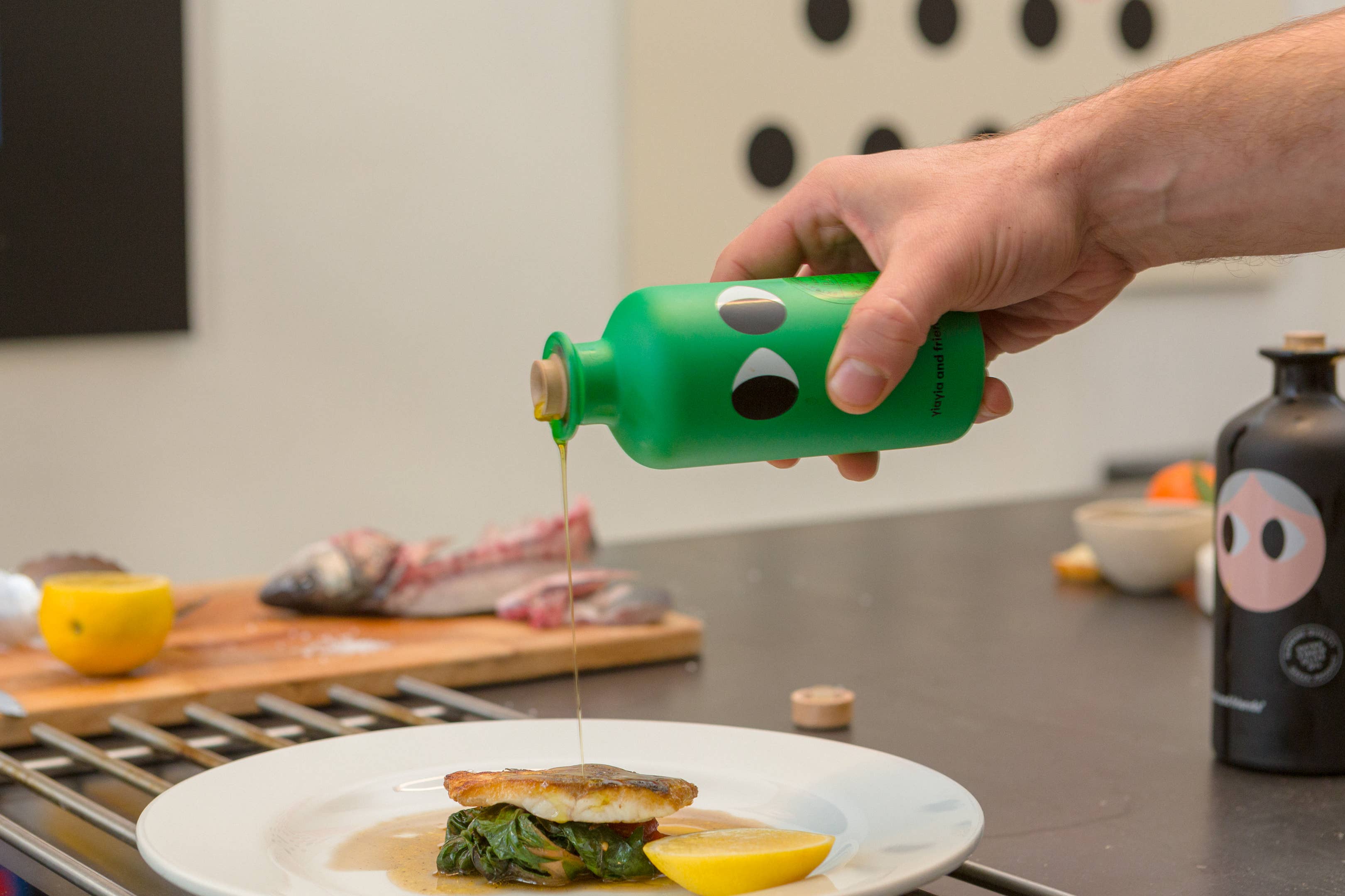 Person pouring a olive oil from a green bottle with cartoon face design onto a plate of food.